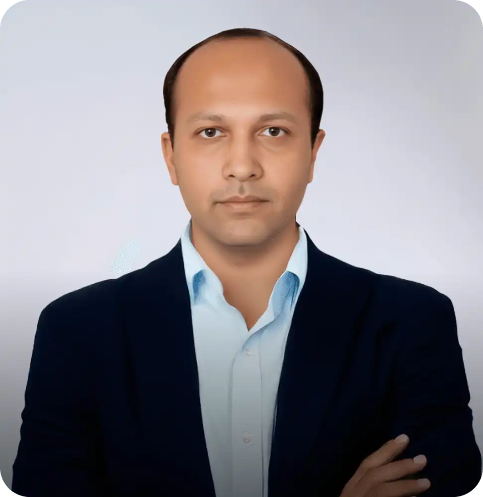 Man in a navy blue suit and light blue shirt posing confidently with folded arms against a plain background.