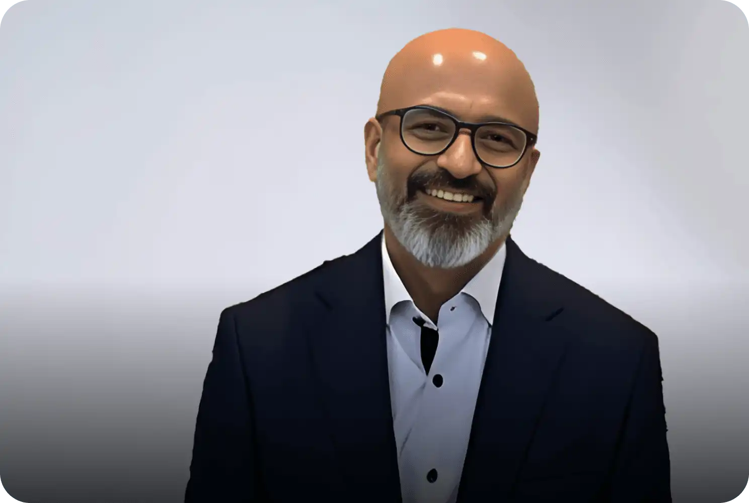 Smiling man with glasses and beard wearing a dark suit and white shirt, posing against a light background.