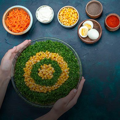 Overhead shot of hands holding a salad with corn and dill arranged in a target shape, surrounded by ingredients.