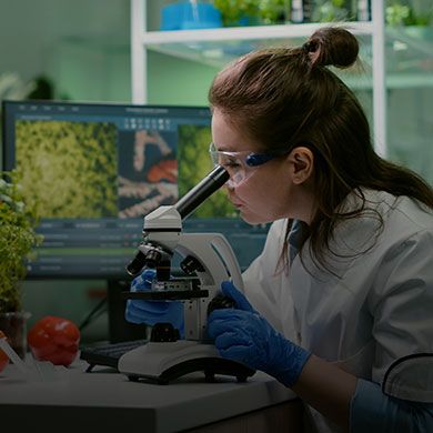 Female biologist in a lab coat and safety glasses intently using a microscope in a research lab.